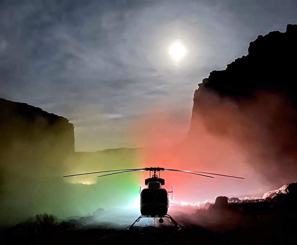 A photo showing the silhouette of a helicopter back-lit by green light to the viewer's left and red light at the viewer's right, with the moon also visible above