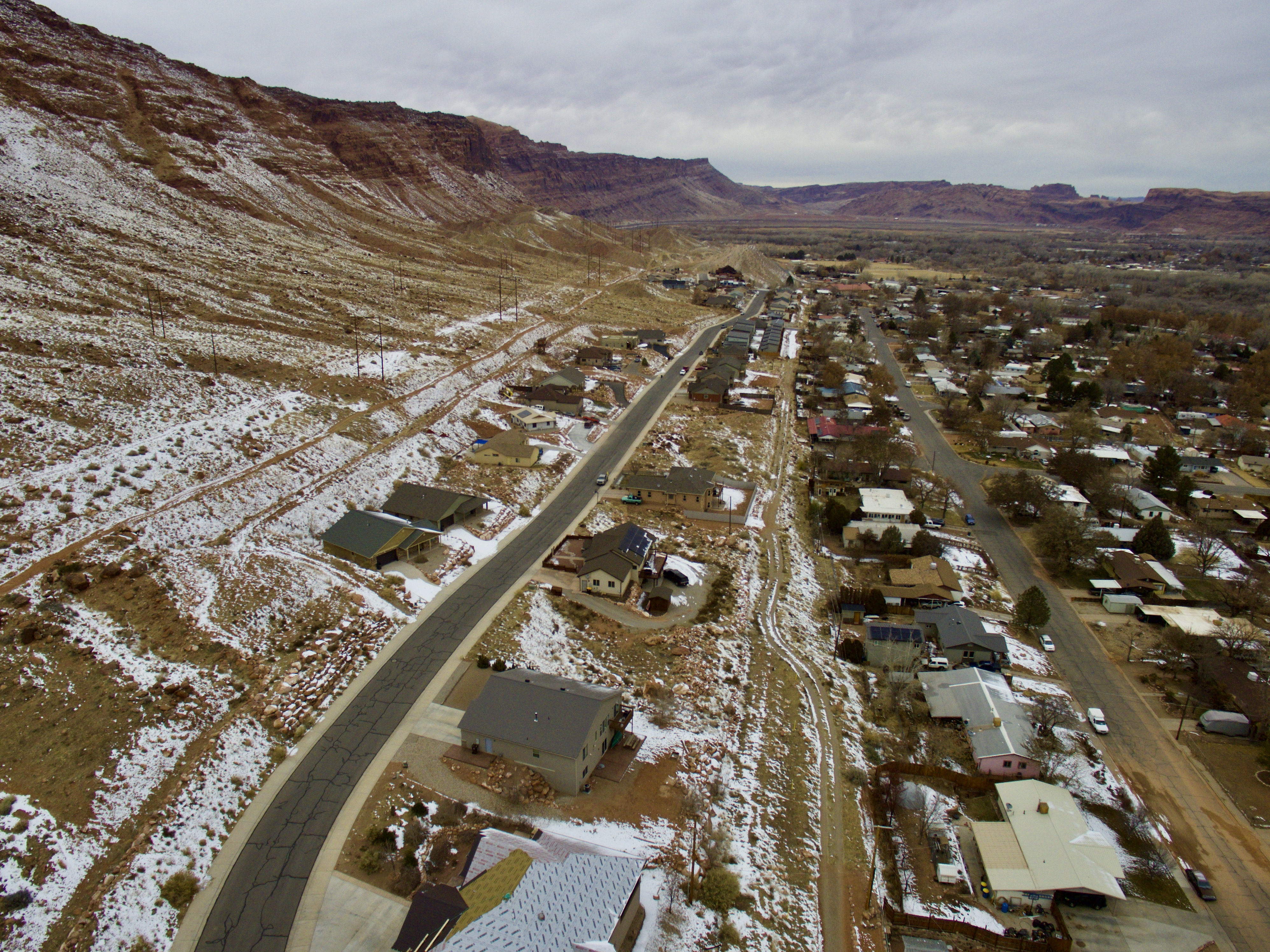 a snowy landscape with homes and, beyond the homes, a sloping area of brush, leading up to a rim of the Moab valley