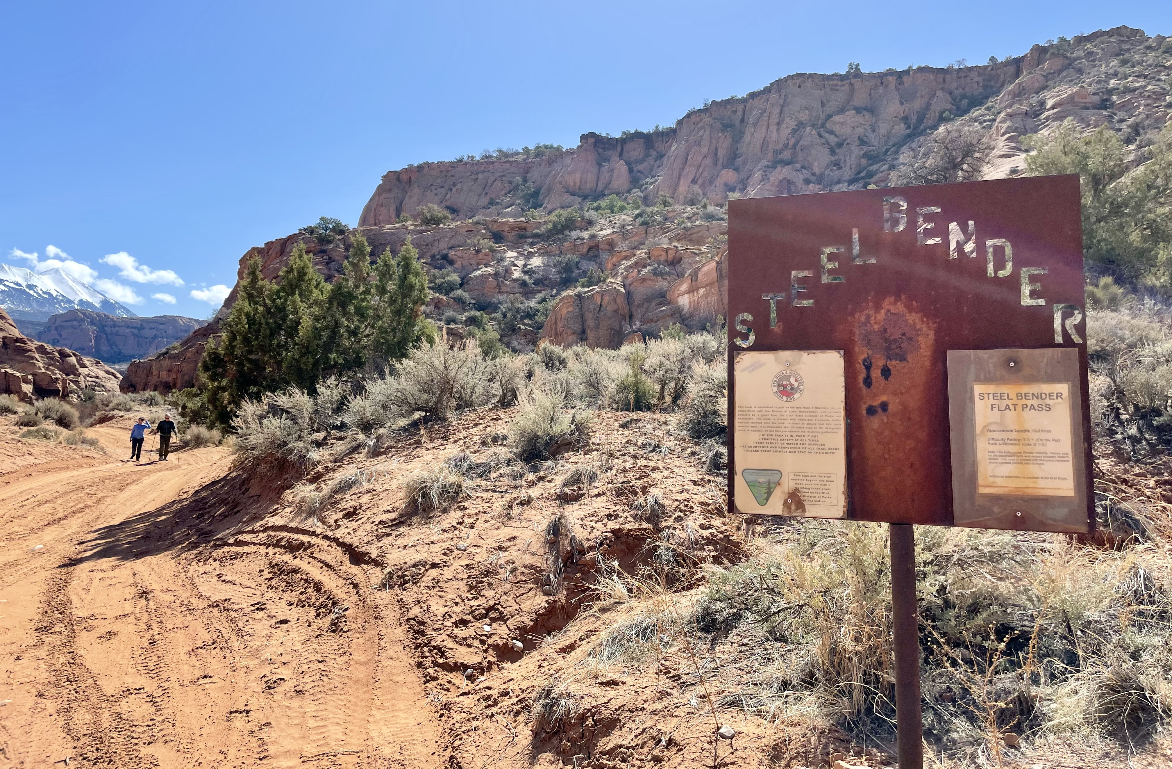 a sign with letters "STEELBENDER" stamped in a chevron pattern displays notices from the Bureau of Land Management about the trail; a couple walks down the trail, toward the viewer, at left