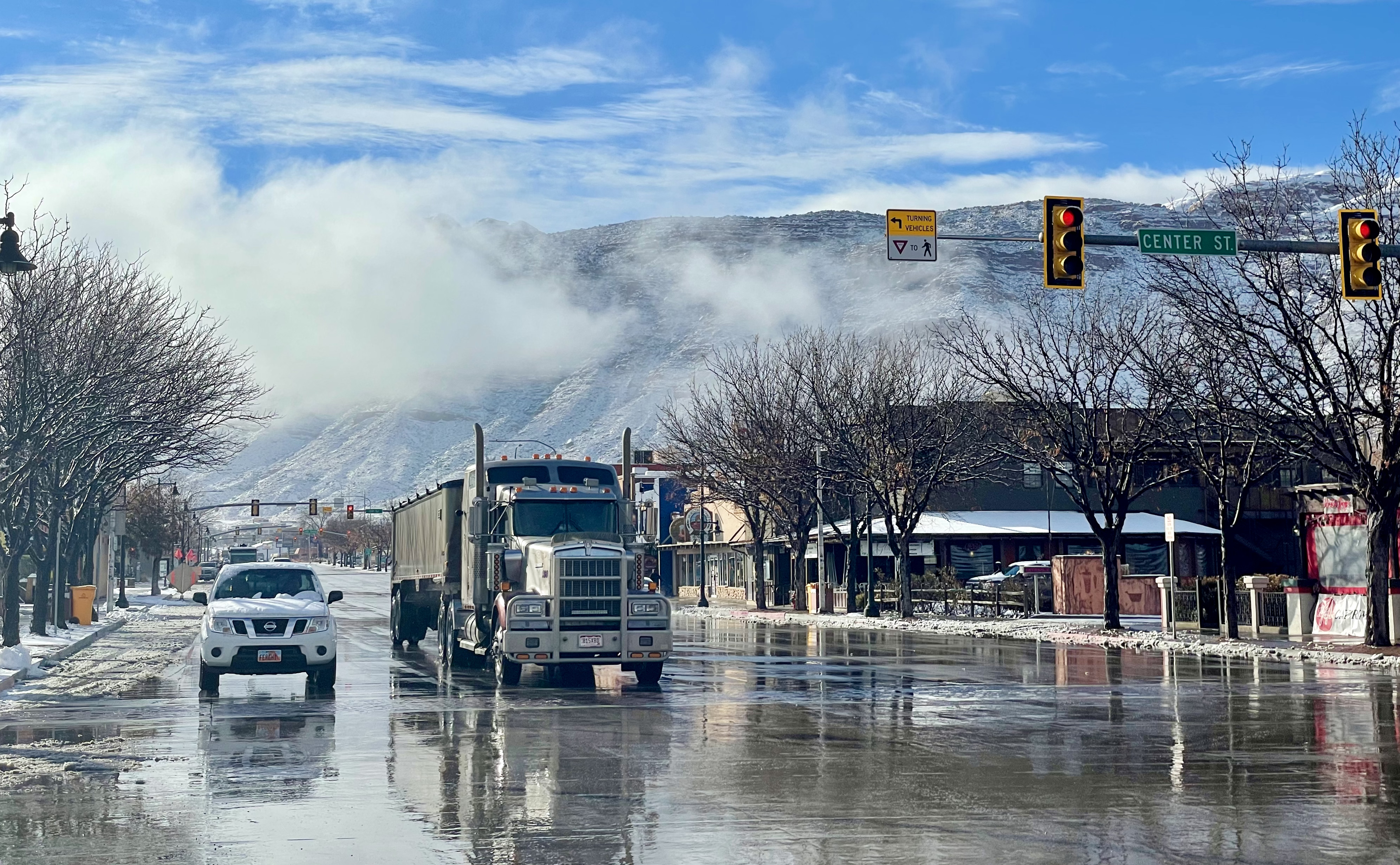 Traffic stops at a red light at a wet intersection; cloud-like haze sits above the trees in the background as the sun shines, melting snow on the ground