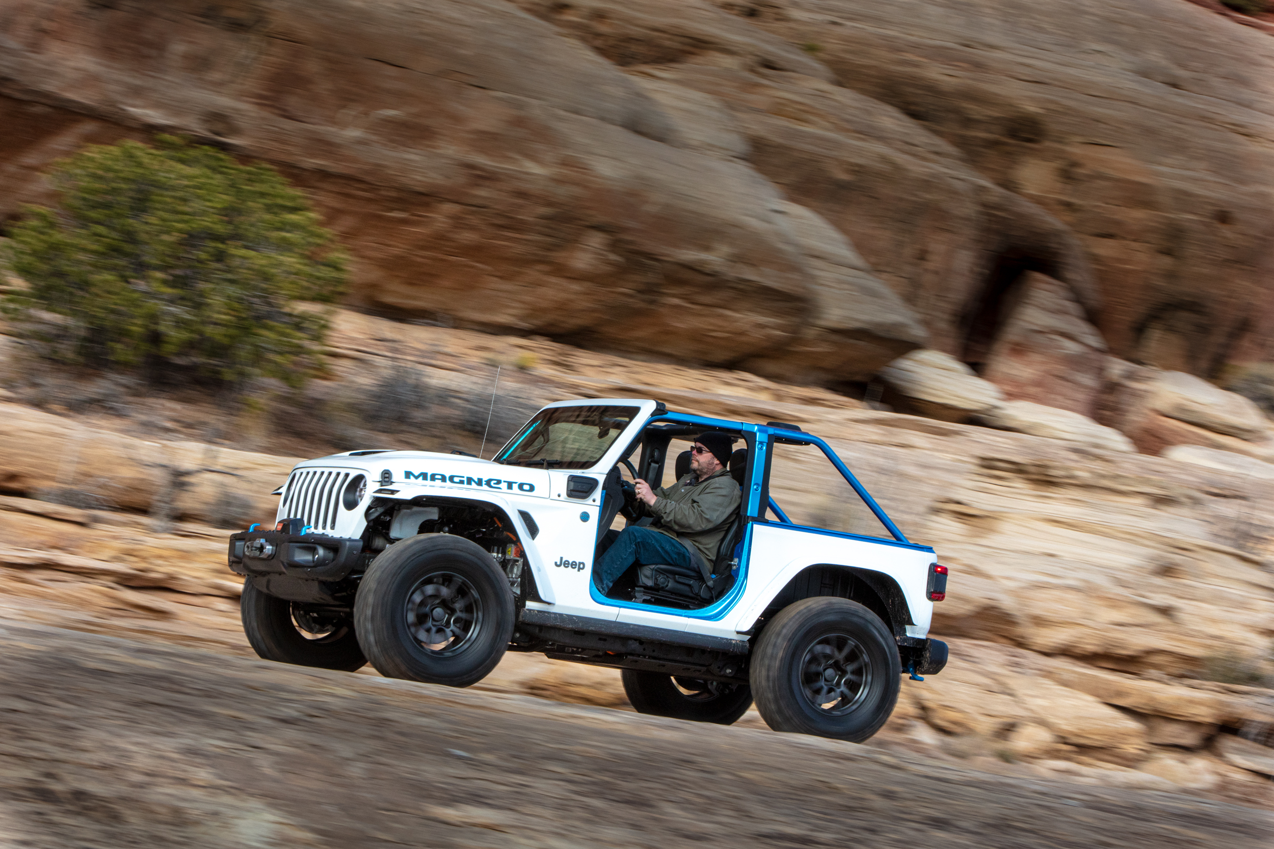 A man drives a white. two-door Jeep with blue trim, and the word Magneto visible on the side of the hood, along smooth slickrock in Moab, Utah.
