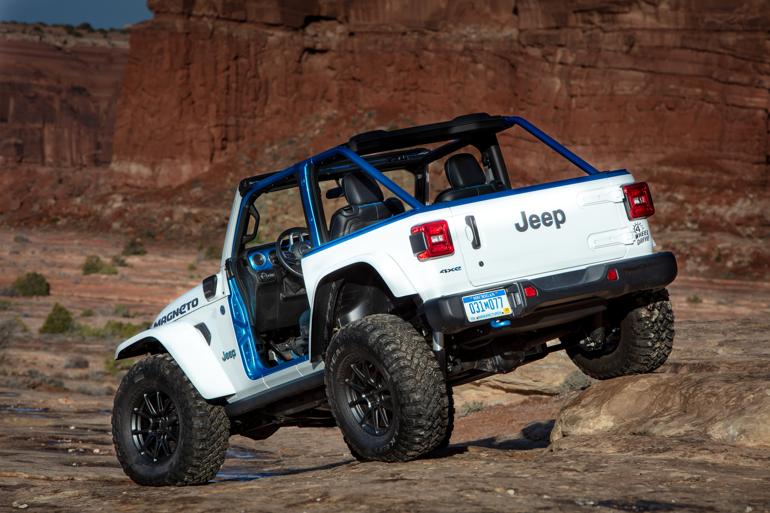 The Jeep sits parked in front of a red cliff, its back right wheel sitting raised on a bulge in the rock underneath.