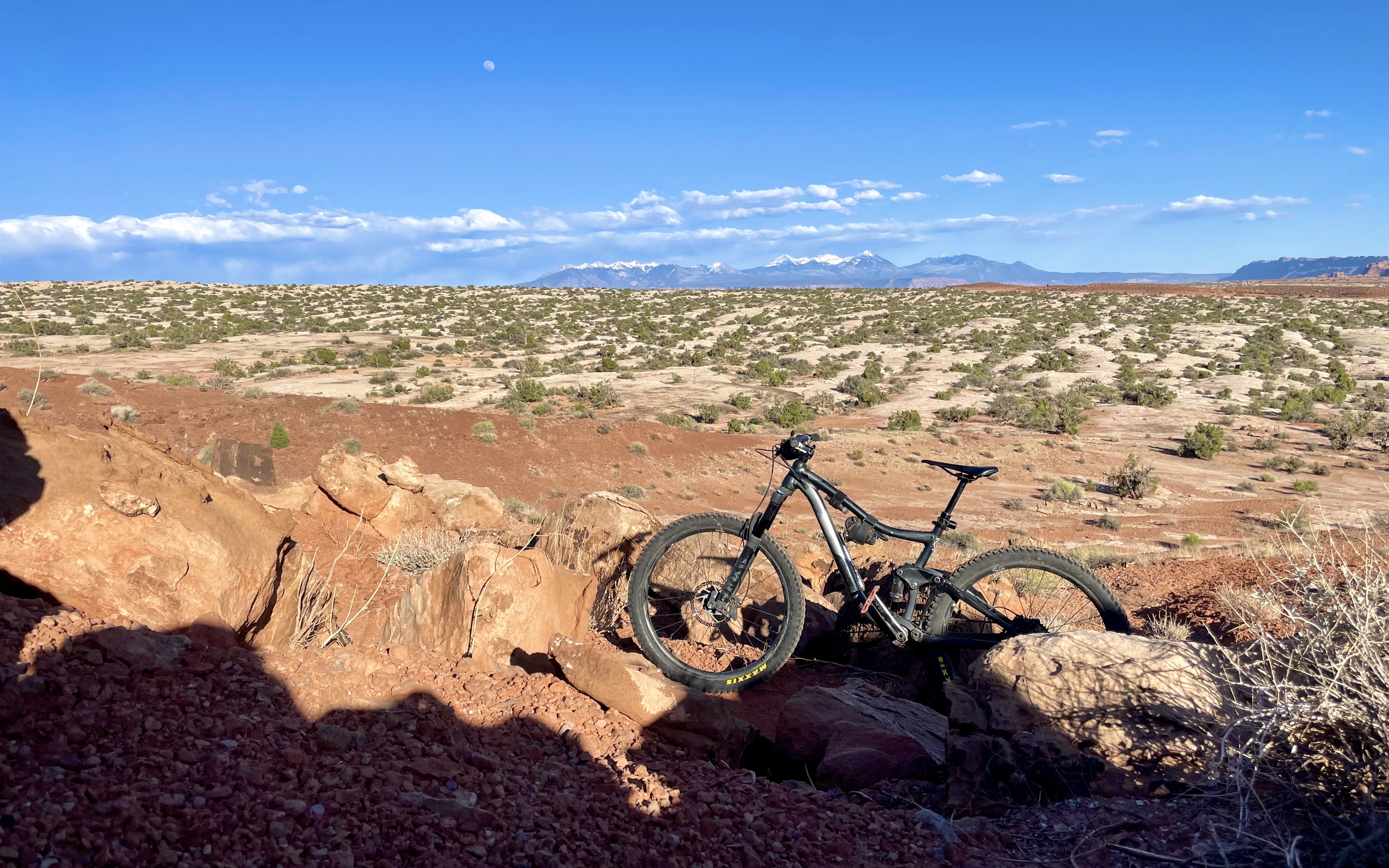 My mountain bike stands in front of a field of slickrock and shrubbery, with the La Sal Mountains and moon in the background